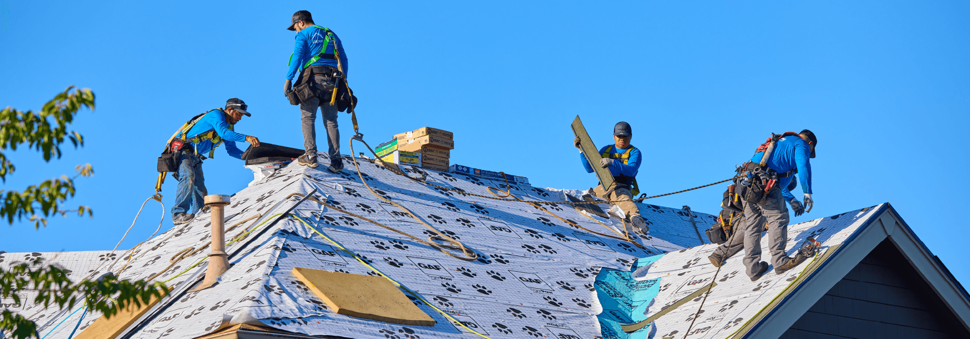 Four workers in safety gear install roofing materials on a steep roof under a clear blue sky.